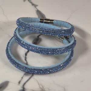 May include: Three light blue bracelets with sparkling blue crystals. The bracelets have a soft, fabric-like texture and a magnetic clasp. The bracelets are stacked on a white marble surface.