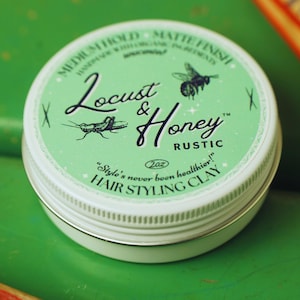 May include: A round, white and green tin of Locust & Honey Rustic Hair Styling Clay. The label features illustrations of a grasshopper and a bee, with the text "MEDIUM HOLD - MATTE FINISH" and "2oz". The product is designed for hair styling.