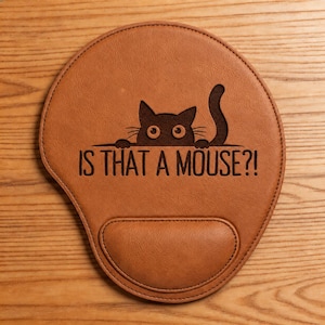 May include: A tan, leather-like mouse pad with a built-in wrist rest. The surface features a dark brown laser-engraved graphic of a cat peeking over a line with the text IS THAT A MOUSE?! positioned directly below the cat illustration.