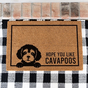 May include: A rectangular tan coir doormat featuring a black border and a black graphic of a Cavapoo puppy peeking over the bottom line. The text HOPE YOU LIKE CAVAPOOS is printed in black capital letters on the right side of the mat.