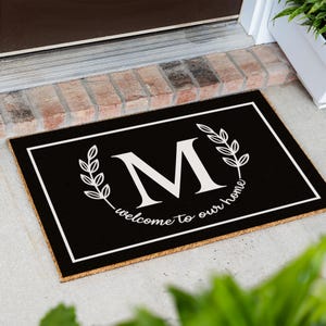 May include: A rectangular black doormat featuring a white border, a large white letter M in the center, and two white laurel branches flanking the letter. Below the letter M, the text "welcome to our home" is printed in white script.