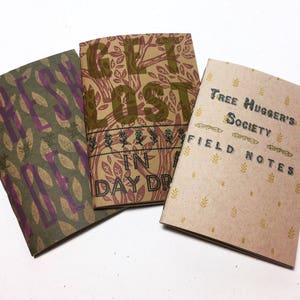 May include: Three handmade notebooks with unique designs. One features the text "FRESH" in purple, another says "GET LOST IN DAY DR" in green, and the third reads "Tree Hugger's Society FIELD NOTES" in green. The notebooks have leaf and floral patterns.
