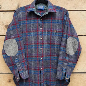 May include: A gray, blue, and red plaid button-down shirt with elbow patches. The shirt has a collared neckline and a single chest pocket. The fabric appears to be a wool blend. The shirt is hanging on a black hanger against a wooden background.