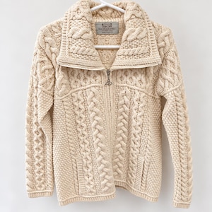 May include: A cream-colored cable knit sweater featuring a full-length front zipper with a metal trinity knot pull. The garment has a wide fold-over collar, long sleeves, and two front pockets, with a label reading Aran Woollen Jacket.