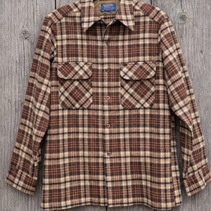 May include: A brown, tan, and navy plaid button-down shirt with two chest pockets. The long-sleeved shirt is hanging on a black hanger against a weathered wood background. The shirt has a collar and a small label.