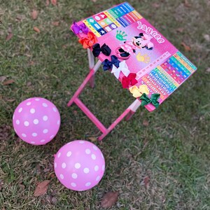 May include: A pink children's table with a Mickey Mouse design and a colourful alphabet chart. The table has a pink and white polka dot balloon on either side. The table is decorated with colourful hair bows.