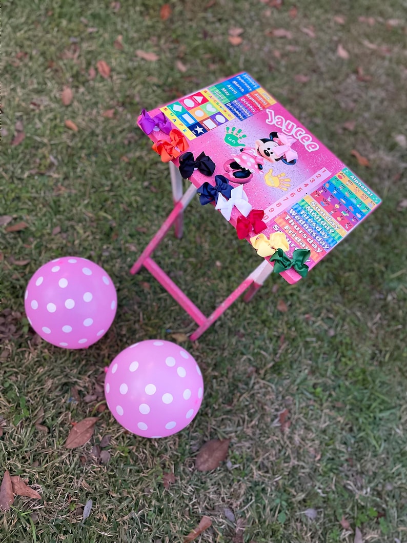 May include: A pink children's table with a Mickey Mouse design and a colourful alphabet chart. The table has a pink and white polka dot balloon on either side. The table is decorated with colourful hair bows.
