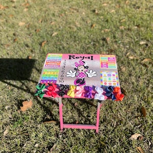 May include: A pink children's table with a Minnie Mouse design and the word "Royal" on the top. The table has a calendar, numbers, and shapes on it. There are colourful bows on the table.