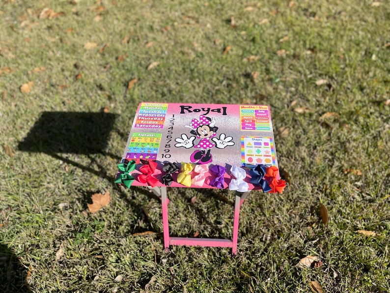 May include: A pink children's table with a Minnie Mouse design and the word "Royal" on the top. The table has a calendar, numbers, and shapes on it. There are colourful bows on the table.