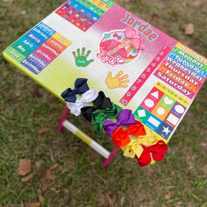 May include: A colourful learning table with a rainbow glitter finish. The table features a calendar, alphabet chart, shapes, numbers, and days of the week. The table also includes a JoJo Siwa sticker and the name Jordae. There are several colourful bows on the table.