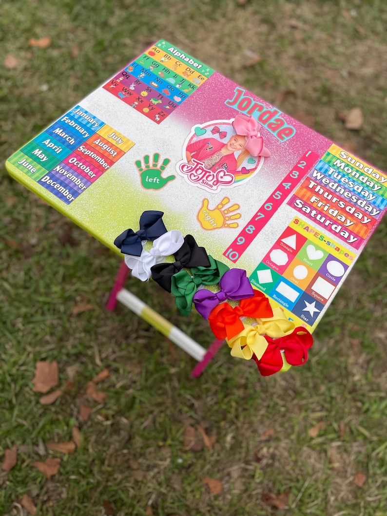 May include: A colourful learning table with a rainbow glitter finish. The table features a calendar, alphabet chart, shapes, numbers, and days of the week. The table also includes a JoJo Siwa sticker and the name Jordae. There are several colourful bows on the table.