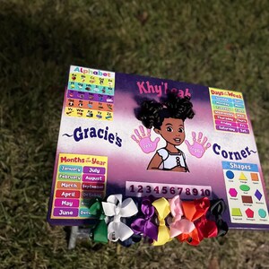 May include: A colourful educational chart with a purple background and a girl's face in the centre. The chart includes an alphabet, days of the week, months of the year, numbers 1-10, and shapes. The chart is decorated with colourful bows.