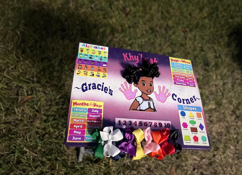May include: A colourful educational chart with a purple background and a girl's face in the centre. The chart includes an alphabet, days of the week, months of the year, numbers 1-10, and shapes. The chart is decorated with colourful bows.