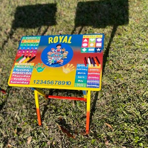 May include: A colourful children's table with a Paw Patrol theme. The table top features an alphabet chart, days of the week, numbers 1-10, shapes, months of the year, and a Paw Patrol logo. The table has a red, yellow, and blue metal frame.