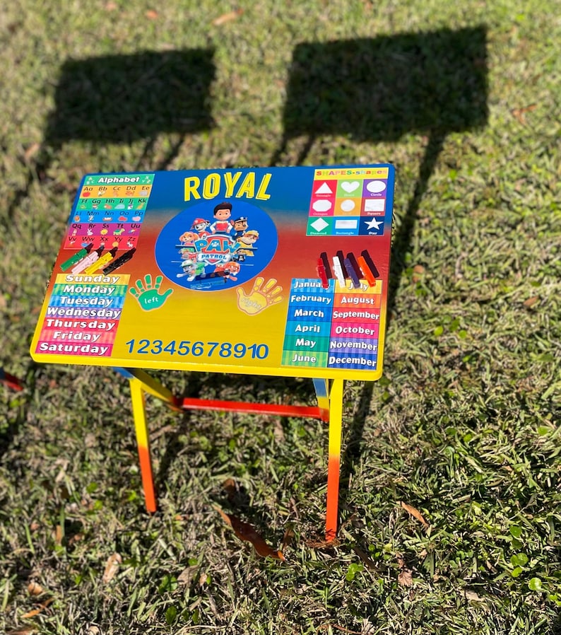 May include: A colourful children's table with a Paw Patrol theme. The table top features an alphabet chart, days of the week, numbers 1-10, shapes, months of the year, and a Paw Patrol logo. The table has a red, yellow, and blue metal frame.