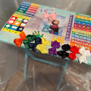 May include: A light blue children's table with a colourful alphabet chart, a calendar, and a picture of Elsa and Anna from Frozen. There are seven colourful bows on the table.