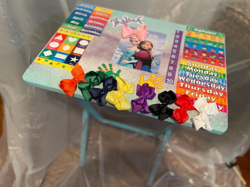 May include: A light blue children's table with a colourful alphabet chart, a calendar, and a picture of Elsa and Anna from Frozen. There are seven colourful bows on the table.