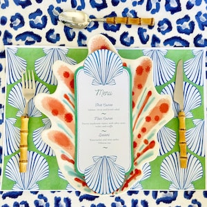 May include: A table setting with a seashell-shaped plate, a menu, and bamboo-handled silverware. The plate is white with coral and teal accents. The menu lists a three-course meal. The background is a green placemat with seashell designs, set on a blue and white leopard print tablecloth.
