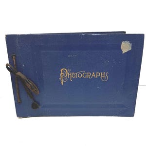 May include: A vintage blue photograph album with the word "Photographs" in gold script. The album has a dark cord tie and a small, peeling sticker on the cover. The album is rectangular and appears to be made of a textured material.