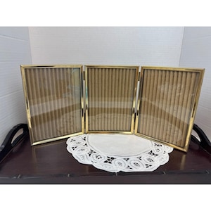 May include: A triptych photo frame featuring three rectangular panels connected by metal hinges. Each frame has a thin, polished gold-tone metal border and contains a clear glass pane over a vertical, ribbed, light brown textured backing.