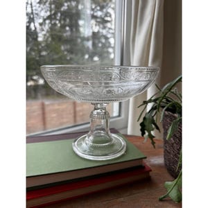 May include: A clear glass pedestal bowl with ornate etched detailing. The bowl sits atop a short, clear stem and a circular base. The bowl is resting on a stack of books with a green cover.