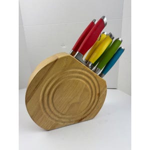 May include: A light-colored wooden knife block with a circular, grooved design on the front. It holds five kitchen knives with colorful handles in shades of red, yellow, lime green, and teal, each featuring a silver-toned metal bolster.