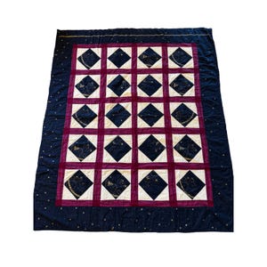 May include: A large, quilted blanket with a repeating pattern of black diamond shapes on a cream background, framed by a burgundy border. The outer border is navy blue with gold star accents.