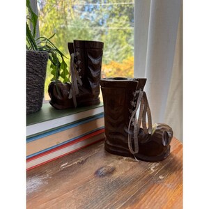 May include: Two dark brown, carved wooden boot-shaped candle holders. Each boot features a glossy finish, a hollow top for a candle, and decorative brown and white checkered fabric laces tied in a bow at the front of the ankle area.