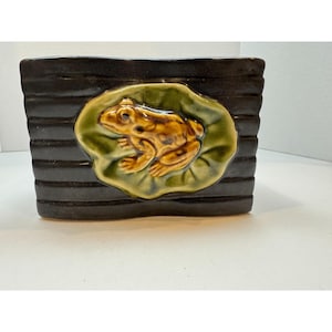 May include: A rectangular, dark brown ceramic planter with horizontal ridges. A raised, detailed frog on a lily pad is centered on the front. The frog is tan and brown, and the lily pad is green.
