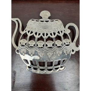 May include: Silver-colored metal teapot-shaped trivet with intricate cut-out seashell and floral designs. The trivet features a handle, spout, and a lid with a seashell finial. This decorative item is designed to protect surfaces from hot cookware.