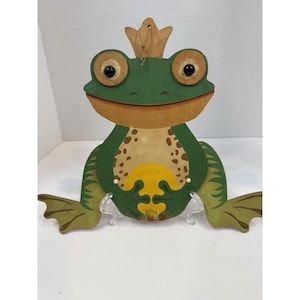May include: Wooden frog figurine with a crown. The frog is painted green with brown spots and a yellow belly. It has large, round eyes and is mounted on a clear stand. The frog's feet are outstretched.