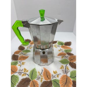 May include: A silver stovetop espresso maker with a bright green handle and top knob. The coffee pot is sitting on a white cloth with a leaf pattern in shades of green, orange, and brown. A classic kitchen appliance.
