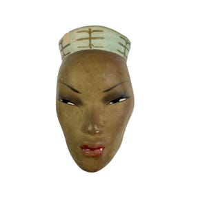May include: A wall-mounted ceramic mask featuring a brown face with almond-shaped eyes, dark eyeliner, and red lips. The mask is topped with a light blue headband decorated with brown, branch-like markings across the forehead area.