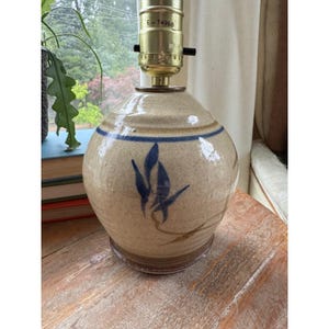 May include: A round, beige ceramic table lamp base with a glossy finish, featuring a hand-painted blue leaf motif and a horizontal blue band near the top. The metal lamp socket at the top displays the text E-74968 and has a dark brown base rim.