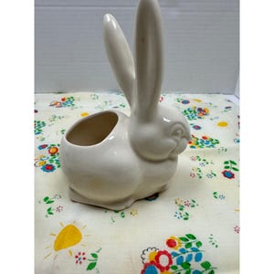 May include: A white ceramic bunny planter with a hollowed-out body for holding small plants or decorations. The bunny has long ears and a detailed face. The planter is sitting on a floral patterned fabric.