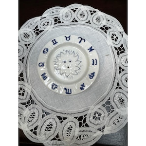 May include: A white ceramic plate with a sun design in the center and zodiac symbols around the edge. The plate is placed on a white lace doily with a detailed, openwork pattern. The overall aesthetic is vintage and decorative.