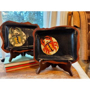 May include: Two decorative trays with crab designs. The trays are black with a red border and sit on wooden stands. One tray features a green and yellow crab, while the other has a red and orange crab.
