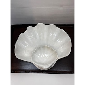 May include: A white, shell-shaped serving dish with a central, smaller dish. The dish has a textured, ridged surface, and is set on a dark brown surface. The design is reminiscent of a clam shell.