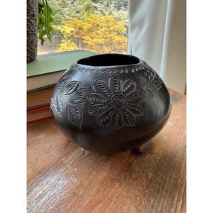 May include: A round, black ceramic vessel featuring hand-carved floral and leaf motifs around its exterior. The surface has a smooth, dark finish with a wide, open circular top and a decorative zigzag pattern etched along the upper rim.