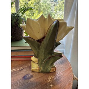 May include: A ceramic vase shaped like a large, blooming flower with pale yellow petals and dark green leaves at the base. The glossy finish highlights the sculpted details of the petals and foliage, creating a dimensional appearance for this decorative piece.