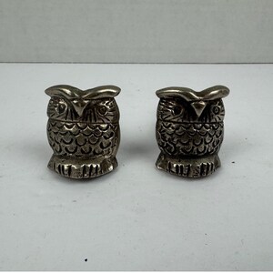 May include: A pair of small, metallic silver-toned owl figurines. Each owl features a textured, scalloped feather pattern on the chest, pointed ear tufts, and a sharp, triangular beak. The figures have a polished, reflective finish and stand upright.
