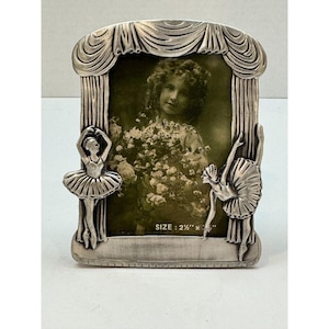 May include: Silver-toned picture frame with a stage curtain design. Two ballerina figures are on either side of the frame. The frame holds a black and white photo. The frame size is 2.5" x 3".