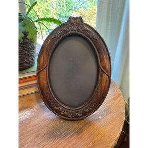 May include: An oval-shaped dark brown wooden picture frame featuring intricate carved floral and leaf motifs along the border. The frame has a smooth, polished finish and a dark, solid-colored insert centered within the oval opening.