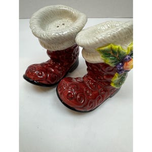 May include: A pair of ceramic salt and pepper shakers shaped like red boots with white fur trim. The boots have a glossy finish and decorative swirl patterns. One boot is decorated with a sprig of holly and berries.