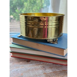 May include: A polished brass planter with a cylindrical shape and horizontal ribbed detailing. The planter features three ornate, curved feet at the base, providing elevation. The metallic surface reflects light, showing a smooth, reflective finish.