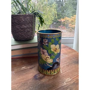 May include: A cylindrical cloisonné vase featuring a vibrant blue background decorated with yellow and white floral patterns, green leaves, and dark rock motifs. The vessel has a gold-toned metal rim and base, with a solid turquoise interior finish.