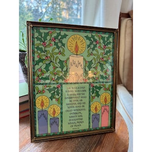 May include: A rectangular brass-framed print featuring a central candle surrounded by green holly leaves and red berries. Below, four smaller candles flank text that reads: For To Us A Child Is Born, To Us A Son Is Given, Isaiah 9:6.