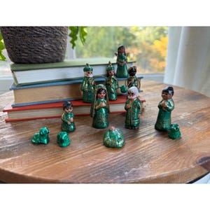 May include: A nativity set featuring small figurines painted in metallic green with gold accents. The collection includes human figures in robes, a small infant in a manger, and several animal figures, all arranged on a wooden surface.