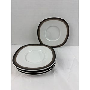 May include: A set of four white ceramic saucers with rounded square shapes. Each saucer features a wide, dark brown band along the outer rim, framing a smooth, white center indentation designed to hold a cup securely in place.