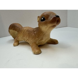 May include: A ceramic squirrel figurine in shades of brown, tan, and cream. The squirrel is posed in a crawling position, with its head tilted upwards. The figurine has detailed fur texture and black eyes.